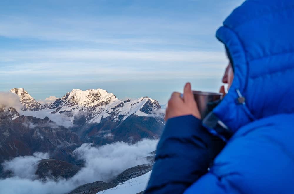 femme alpinite buvant un café au sommet d'une montagne avec une doudoune a capuche bleue